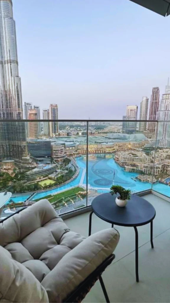 High-angle view from a modern luxury apartment balcony overlooking a vibrant metropolitan center at dusk. A comfortable, deeply cushioned grey outdoor chair faces a glass panel balustrade next to a small black accent table holding a tiny plant. Below, a massive, uniquely shaped bright blue outdoor water feature twists through a high-end commercial complex, surrounded by towering glass skyscrapers reaching into a clear sky.