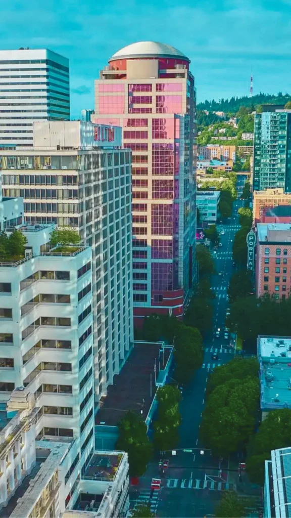 An aerial, high-angle shot looking down a straight city avenue in downtown Portland, bordered by green street trees and high-rise buildings. On the left, a tall white concrete structure with small rooftop terraces stands adjacent to a unique, tall skyscraper featuring reflective pink and red glass panels and a domed roof. In the background, a historic marquee sign reading "PORTLAND" is visible over the street, with a forested hill stretching under a blue sky.
