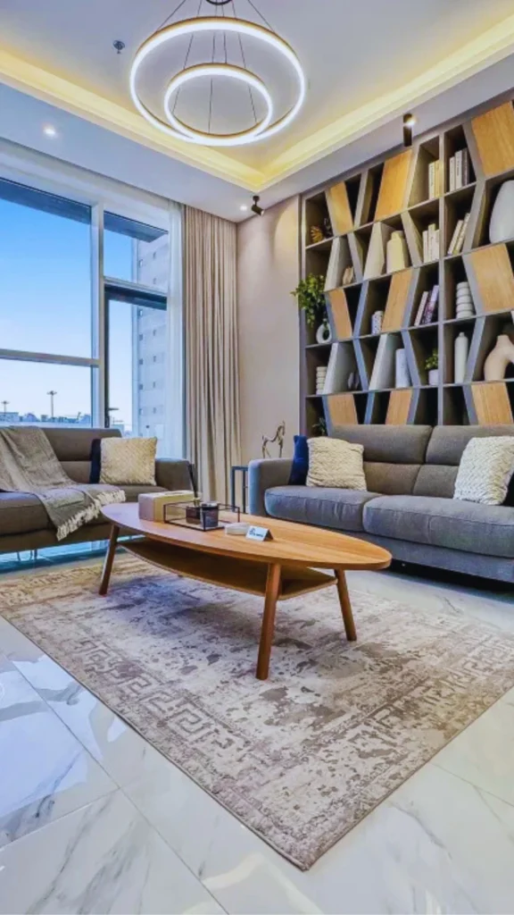 Interior view of a high-end contemporary apartment living space. The room includes a long charcoal grey sofa, a smaller coordinating loveseat, and an oval tiered wooden coffee table resting on an patterned light brown area rug. In the background, an intricate floor-to-ceiling display cabinet with diagonal wooden divider panels holds books and pottery, while a large glass window wall opens up to bright daylight.
