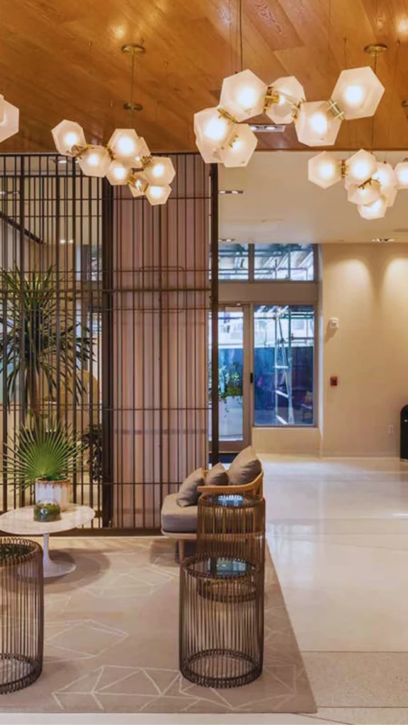 Interior view of a contemporary luxury hotel lobby or apartment building common space. In the foreground, cylindrical metal wire-frame side tables sit on a geometric-patterned beige rug next to a modern sofa with cushions. A tall metal grille room divider separates the lounge from large glass exit doors in the background, all illuminated by striking, frosted hexagonal link light fixtures hanging from a natural wood ceiling.