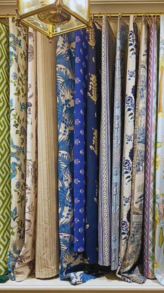 A luxury showroom display highlighting an array of premium curtain fabrics that add depth and a palpable richness to interior design. A series of heavy, high-end drapery panels hang closely together from a classic gold brass curtain rod. The collection features sophisticated textures and rich tones, including deep jewel-blue printed silks, intricate botanical patterns, a neutral-toned raw silk panel, and bold geometric green textiles, all softly illuminated by a vintage gold pendant lantern hanging in the foreground.
