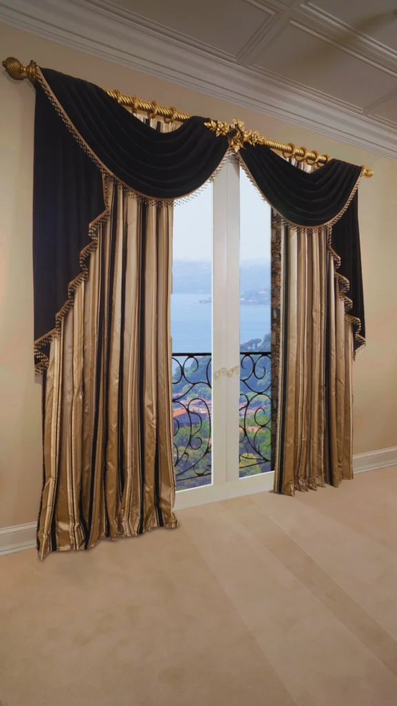 An elegant pair of floor-length curtains surrounding a set of white French doors that open up to a coastal view. The main curtain panels feature a vertical striped pattern in shimmering gold and dark charcoal black. Draped across the top of the window is a dramatic, deep black velvet valance adorned with gold tasselled trim along its curved edges. The entire window treatment hangs from a decorative, twisted brass rod featuring ornate gold leaf centerpieces and end finials.