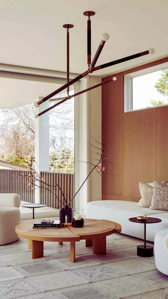 Interior view of a contemporary, upscale living room displaying organic furniture designs. A low, rounded white fabric sofa with textured bouclé accent pillows faces a low-profile circular wooden coffee table decorated with books and a vase holding minimal flowering branches. Overhead hangs a large, minimalist black and brass linear pendant light fixture with round globe bulbs, emphasizing sculptural flow against a wood accent wall and large floor-to-ceiling windows.