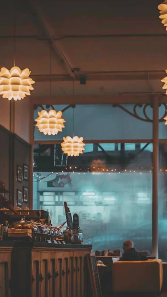 An interior space illustrating the visual contrast between distinct warm and cool lighting zones. Indoors, a row of decorative, multi-layered artichoke-style pendant lamps casts a dim, cozy amber glow over a service counter and seating area, creating a relaxing evening or winding-down ambiance. In sharp contrast, a large window reveals a cool, bright blue-toned environment outside, demonstrating how different light spectrums can visually separate zones.